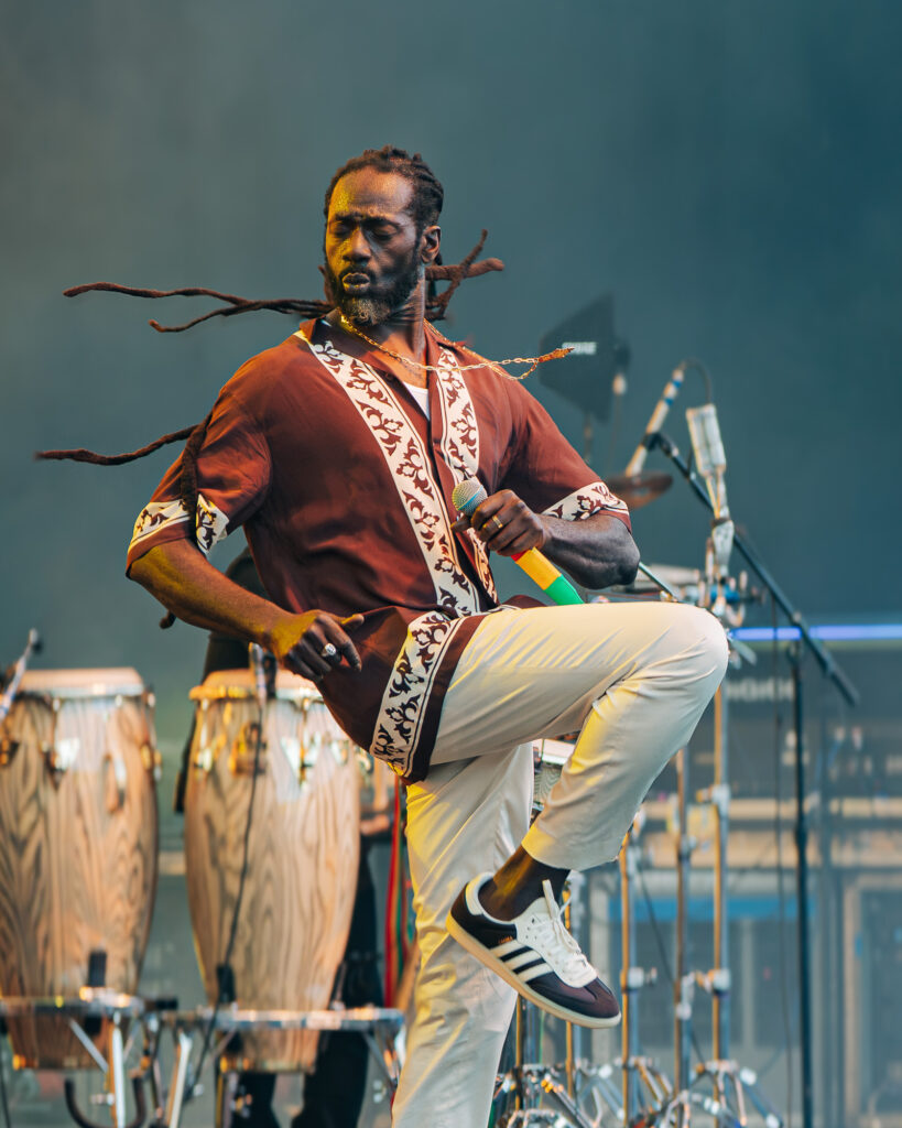 Buju-Banton-Roots-and-Reggae-Concert-Pacific-Amphitheatre-OC-June-19-2026- – The 824 Journal Male musician with dreadlocks in a brown patterned shirt, mid-performance on stage beside conga drums, holding a microphone.