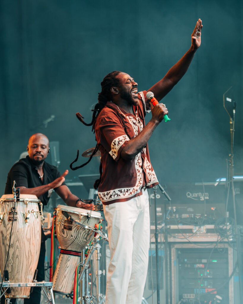 Buju-Banton-Roots-and-Reggae-Concert-Pacific-Amphitheatre-OC-June-19-2026-2026046 – The 824 Journal Male singer on stage, microphone in hand, arm raised in performance with a drummer and percussion instruments nearby.
