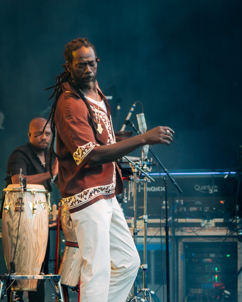 Buju-Banton-Roots-and-Reggae-Concert-Pacific-Amphitheatre-OC-June-19-2026-2026108 – The 824 Journal Male performer with dreadlocks on a dim stage, wearing a maroon shirt and white pants, singing or playing to a crowd; congas visible on the left.