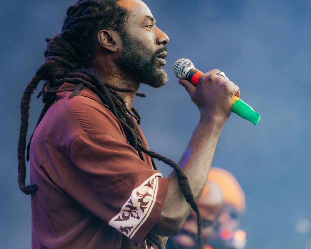 Buju-Banton-Roots-and-Reggae-Concert-Pacific-Amphitheatre-OC-June-19-2026--3 – The 824 Journal Male singer with long dreadlocks holding a microphone close to his mouth during a live performance on a blue-lit stage.