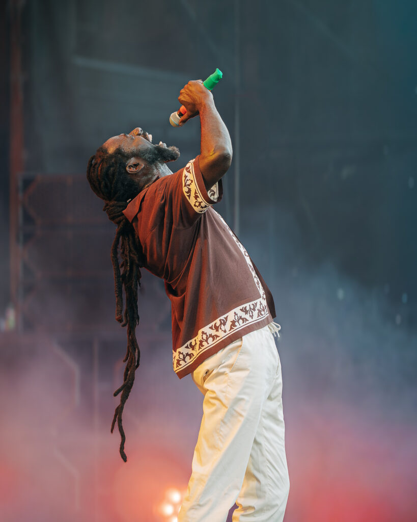 Buju-Banton-Roots-and-Reggae-Concert-Pacific-Amphitheatre-OC-June-19-2026--4_(2) – The 824 Journal Male performer singing into a microphone on stage, wearing a brown patterned shirt with white trim and long dreadlocks framing his face.