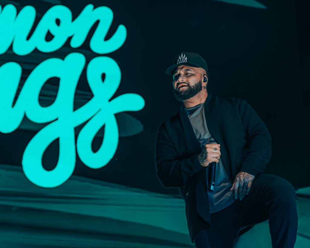 Common-Kings-Concert-Los-Angeles-May-23-2026-by-Zharmaaaine-68 – The 824 Journal Male performer kneels on a dark stage, microphone in hand, wearing a cap with a crown logo, with large teal neon letters in the background behind him.