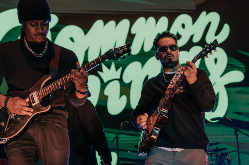 Common-Kings-Concert-Los-Angeles-May-23-2026-by-Zharmaaaine-73 – The 824 Journal Two guitarists performing on a dimly lit stage with a bright green neon backdrop; one wears a beanie and necklace, the other sunglasses and patterned strap on his guitar.