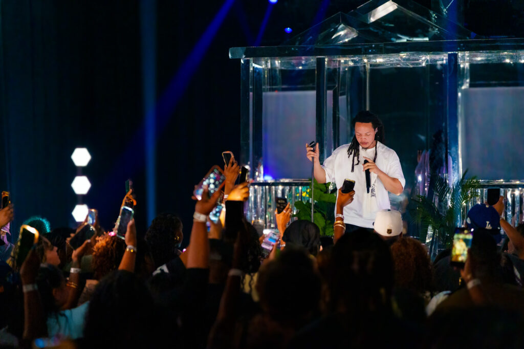 Domani-The-Echo-Los-Angeles-June-23-2026-by-Zharmaine-Boatman-IG-zharmaaaine-15-2 – The 824 Journal Male performer with dreadlocks in a white shirt stands inside a glass booth on stage, holding a microphone as lights glow and fans raise phones in the crowd.
