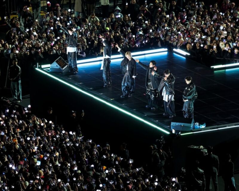 A band performing on an elevated stage with blue neon edges, as a large crowd waves phone lights in front of them.