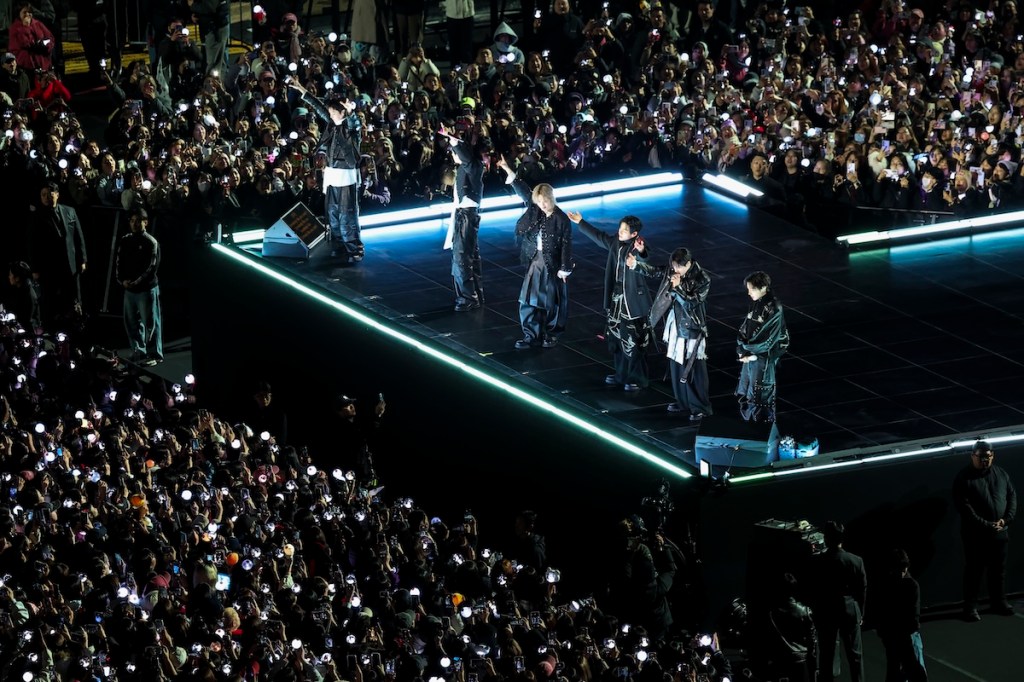 A band performing on an elevated stage with blue neon edges, as a large crowd waves phone lights in front of them.