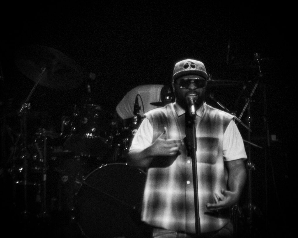Musiq-Soulchild-The-Grove-of-Anaheim-May-29-2026-16 – The 824 Journal Male singer on stage, wearing a skull-cap and sunglasses, singing into a microphone with a plaid shirt, drums in the background (black-and-white).