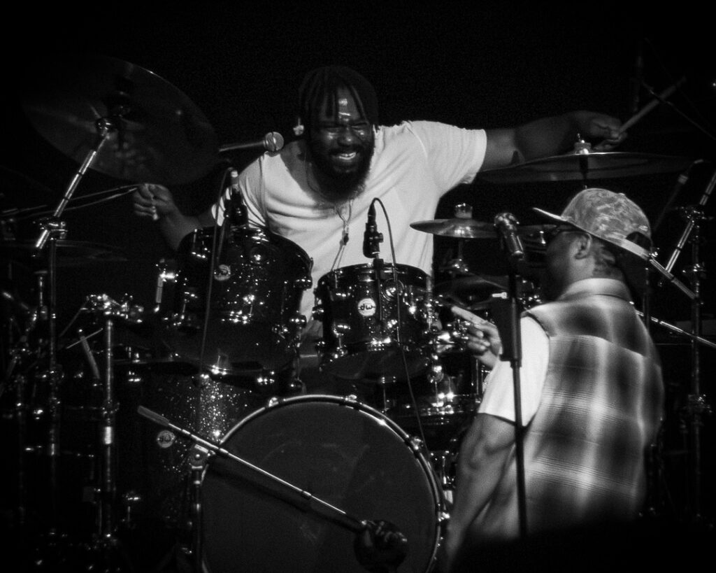 Musiq-Soulchild-The-Grove-of-Anaheim-May-29-2026-18 – The 824 Journal Bearded drummer smiling behind a sparkling drum kit on a dark stage, while another musician faces away, wearing a cap and checkered vest.