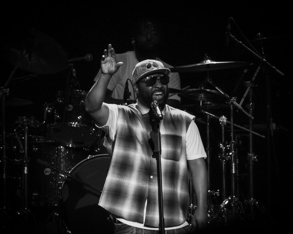 Musiq-Soulchild-The-Grove-of-Anaheim-May-29-2026-19 – The 824 Journal Male singer on stage, wearing cap and sunglasses, raising his left hand as he performs; drummer in the background.
