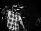 Male performer on stage in a plaid shirt, cap and sunglasses points to audience with a smile while a drummer and drums are visible in the background.