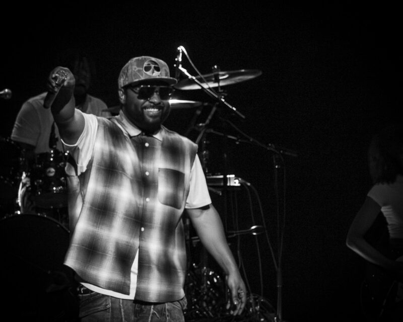 Male performer on stage in a plaid shirt, cap and sunglasses points to audience with a smile while a drummer and drums are visible in the background.