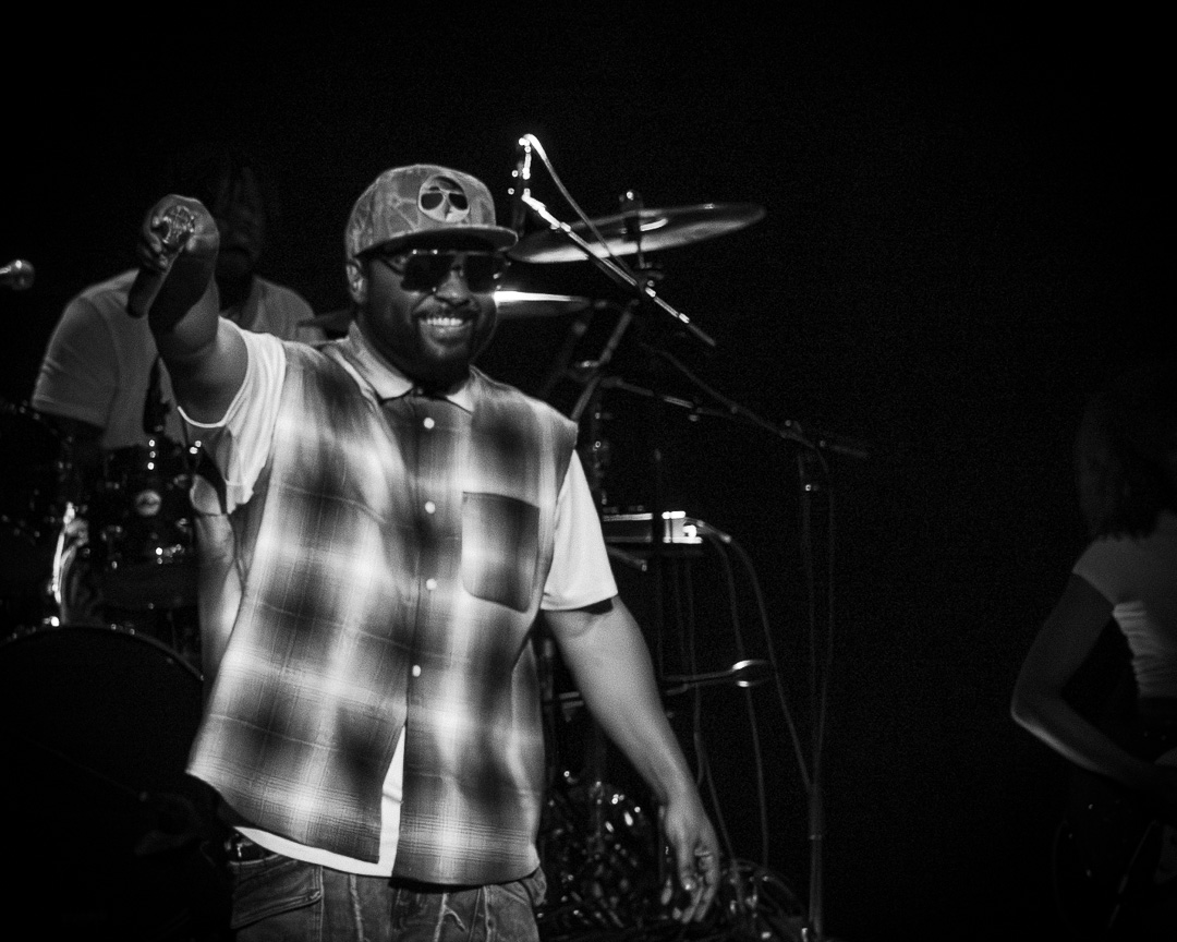 Male performer on stage in a plaid shirt, cap and sunglasses points to audience with a smile while a drummer and drums are visible in the background.
