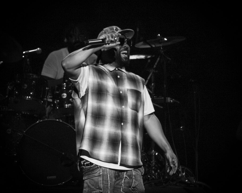 Musiq-Soulchild-The-Grove-of-Anaheim-May-29-2026-28 – The 824 Journal Man singing into a microphone on stage, wearing a plaid shirt and cap, with a drummer and drum kit in the background (black and white).**