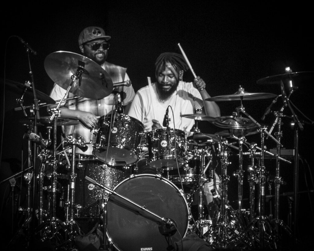 Musiq-Soulchild-The-Grove-of-Anaheim-May-29-2026-44 – The 824 Journal Two male drummers on stage playing a shiny drum kit; one raises a drumstick with a smile behind the kit.