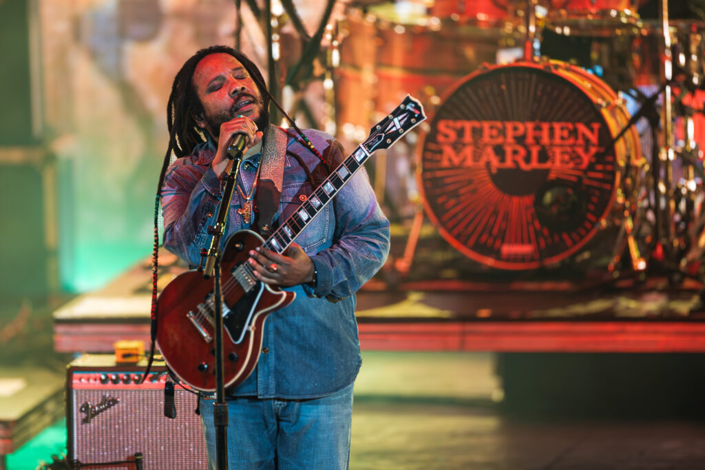 Stephen-Marley-Roots-and-Reggae-Concert-Pacific-Amphitheatre-OC-June-19-2026- – The 824 Journal Musician with dreadlocks sings into a microphone while playing a guitar on stage, with a large Stephen Marley drum backdrop behind him.