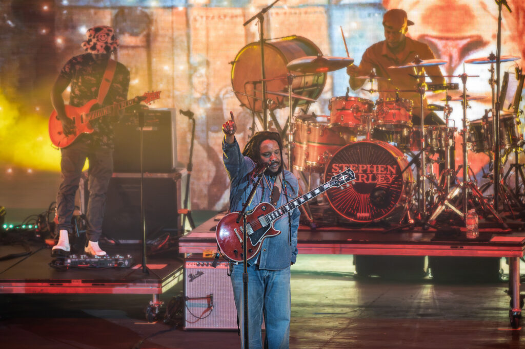 Stephen-Marley-Roots-and-Reggae-Concert-Pacific-Amphitheatre-OC-June-19-2026--2 – The 824 Journal Live concert scene with a guitarist front and center, singer raising his fist, drummer behind, and colorful stage lights in the background.
