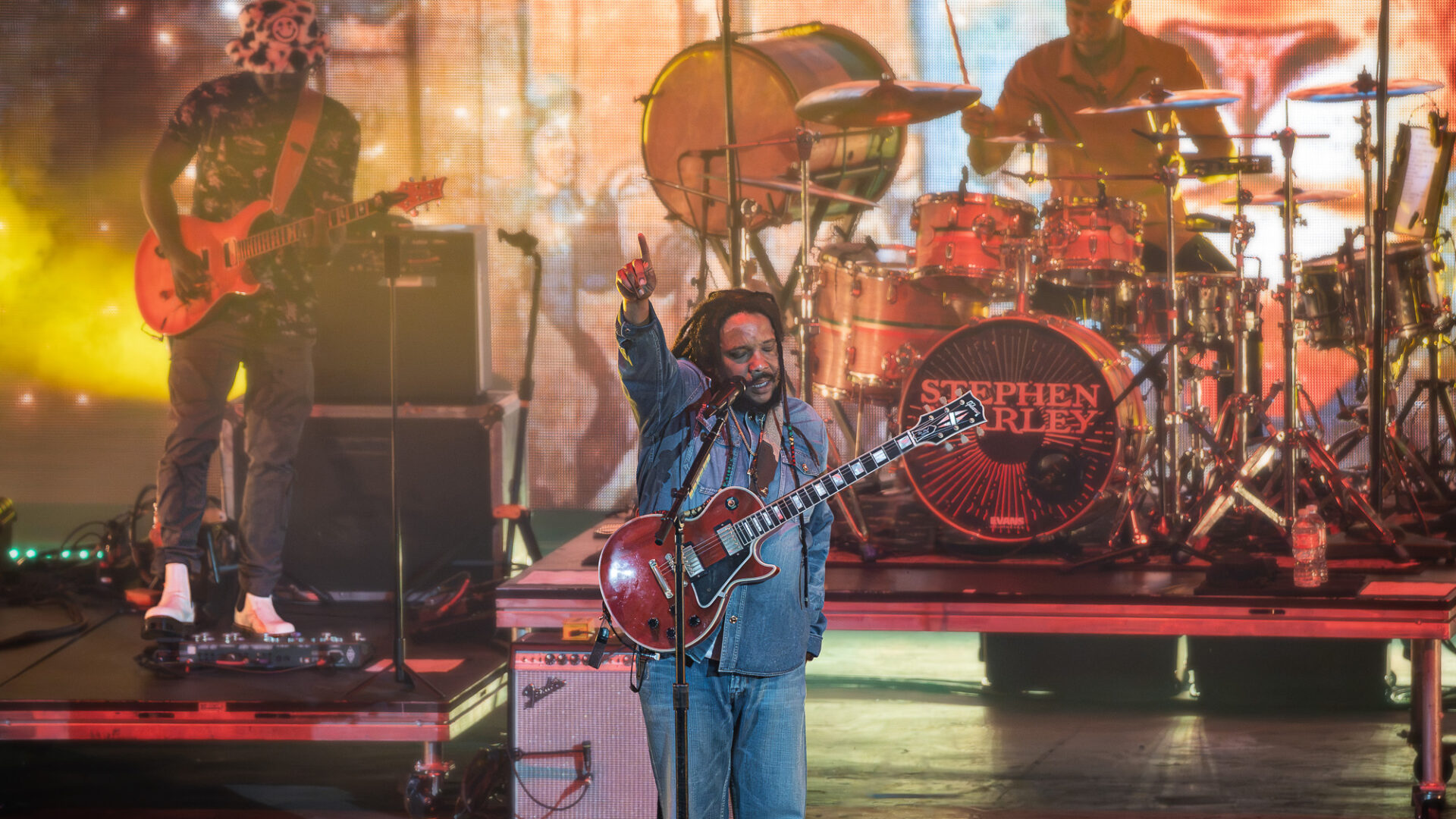 Stephen-Marley-Roots-and-Reggae-Concert-Pacific-Amphitheatre-OC-June-19-2026--2 – The 824 Journal Live concert scene with a guitarist front and center, singer raising his fist, drummer behind, and colorful stage lights in the background.