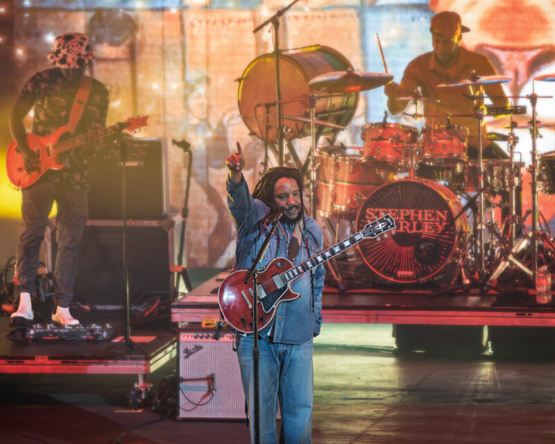 Live concert scene with a guitarist front and center, singer raising his fist, drummer behind, and colorful stage lights in the background.