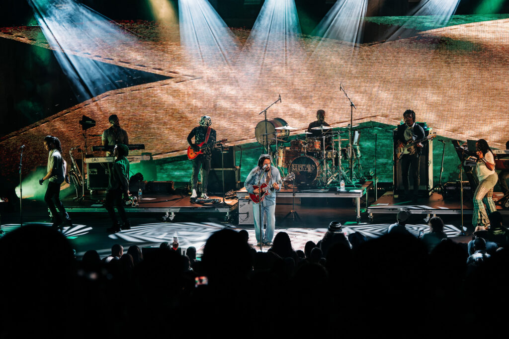 Stephen-Marley-Roots-and-Reggae-Concert-Pacific-Amphitheatre-OC-June-19-2026-20261206 – The 824 Journal Live reggae band on stage performing to an audience with multiple musicians, guitars, keyboards, drums, and backlit backdrop projection behind them.