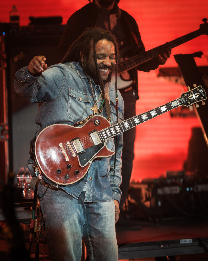 Stephen-Marley-Roots-and-Reggae-Concert-Pacific-Amphitheatre-OC-June-19-2026-20261244 – The 824 Journal Guitarist with dreadlocks in a denim shirt smiling while playing a sunburst Gibson Les Paul on a stage with red lighting.