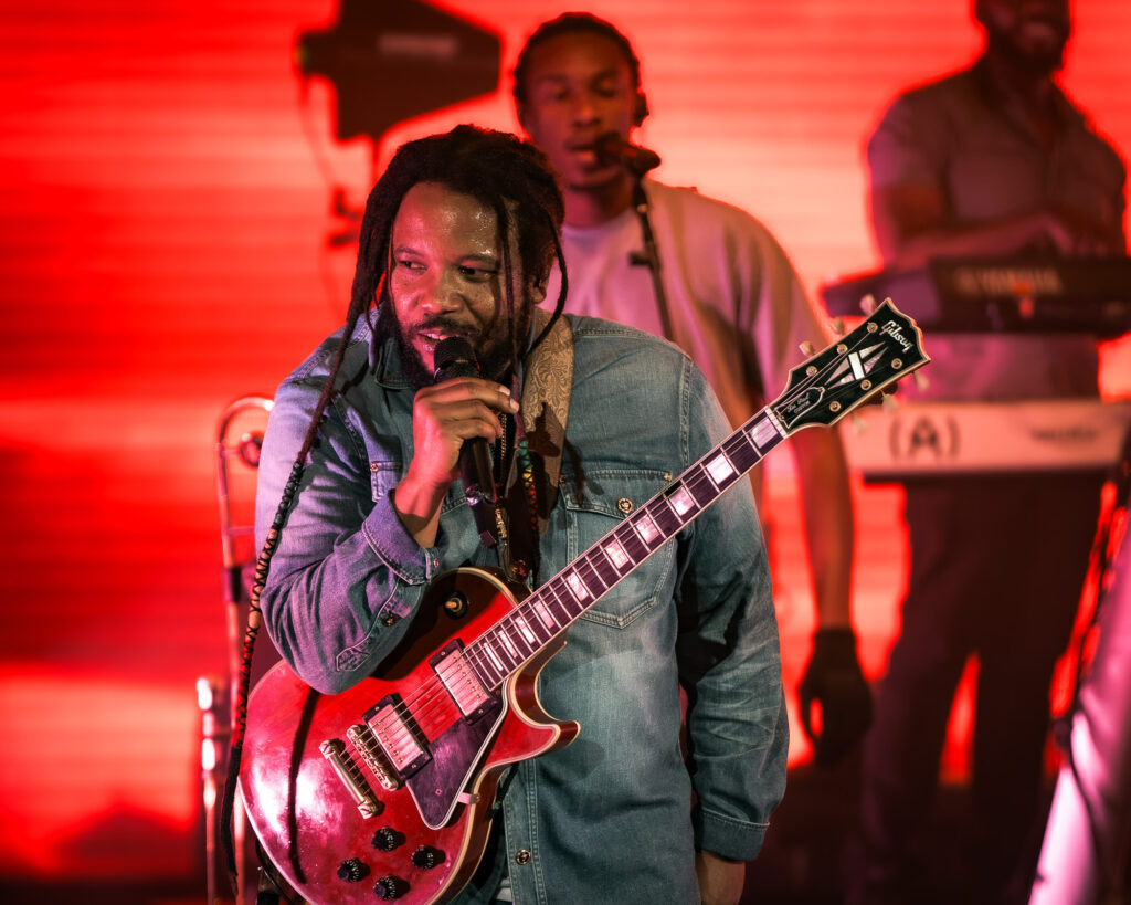 Stephen-Marley-Roots-and-Reggae-Concert-Pacific-Amphitheatre-OC-June-19-2026-20261352 – The 824 Journal Male guitarist sings into a microphone on stage, wearing a denim jacket, with a red-lit backdrop and a keyboardist in the background.