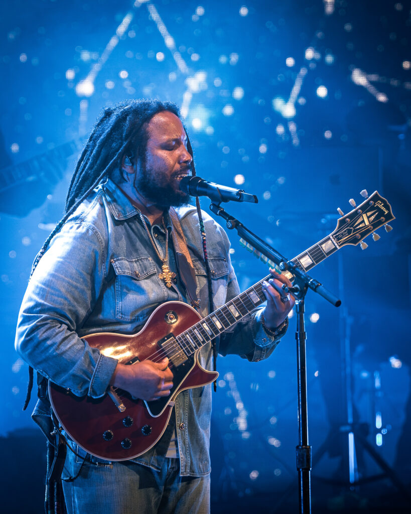 Stephen-Marley-Roots-and-Reggae-Concert-Pacific-Amphitheatre-OC-June-19-2026-20261451 – The 824 Journal Male guitarist with dreadlocks, denim jacket, singing into a microphone on a blue-lit stage with a red guitar.