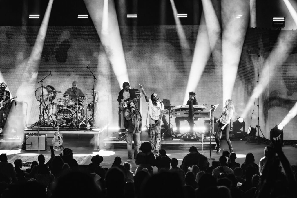 Stephen-Marley-Roots-and-Reggae-Concert-Pacific-Amphitheatre-OC-June-19-2026-20262106 – The 824 Journal Live concert scene with a band performing on stage under bright spotlights, singer at center front, guitarist and drummer visible, and a cheering audience in the foreground.