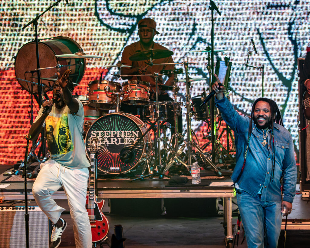 Stephen-Marley-Roots-and-Reggae-Concert-Pacific-Amphitheatre-OC-June-19-2026--3 – The 824 Journal Singer front left with microphone on a brightly lit stage, drummer Stephen Marley in the center behind a drum kit, guitarist at left, and another performer in denim on the right; colorful LED backdrop fills the scene.