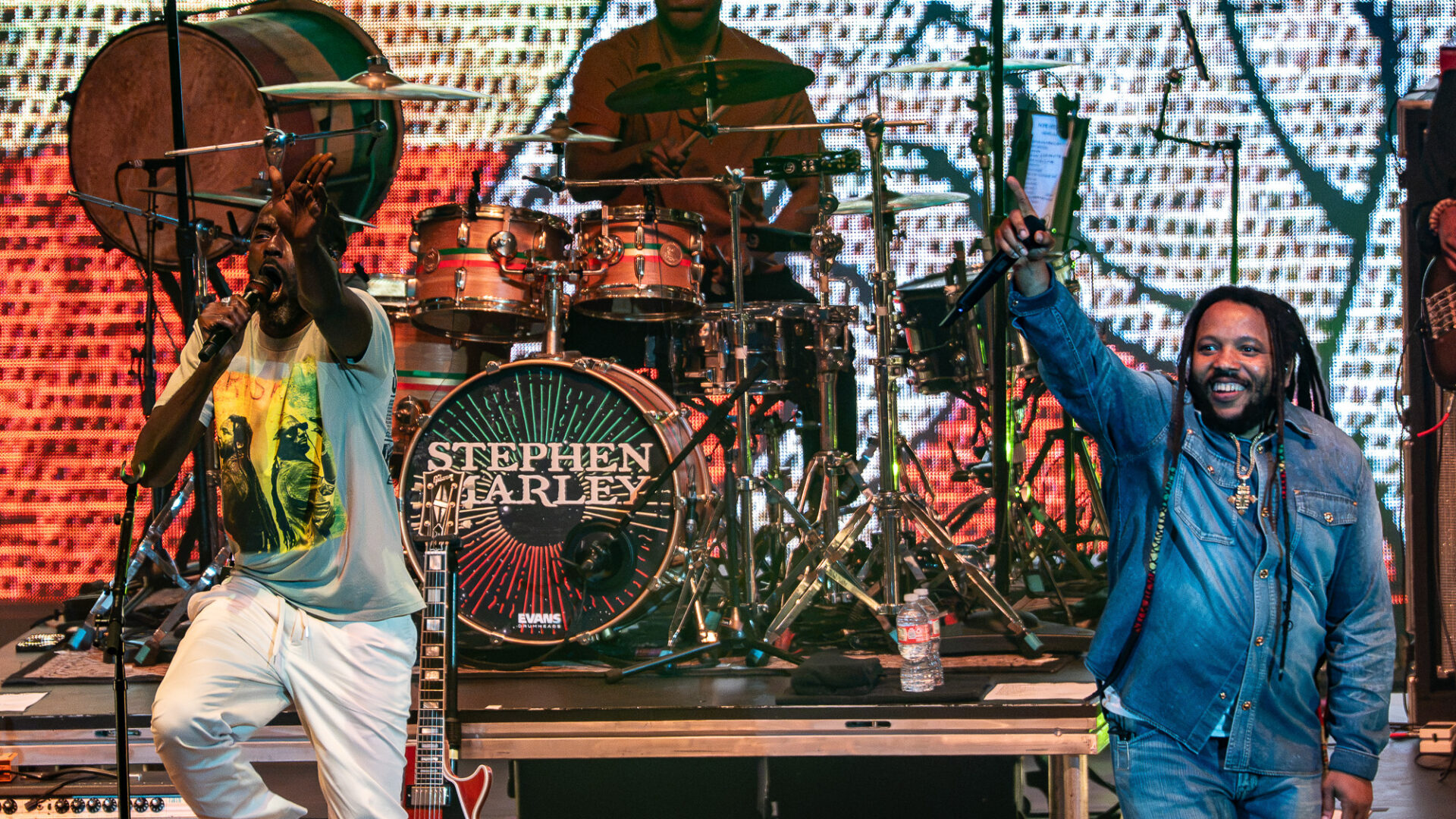 Stephen-Marley-Roots-and-Reggae-Concert-Pacific-Amphitheatre-OC-June-19-2026--3 – The 824 Journal Singer front left with microphone on a brightly lit stage, drummer Stephen Marley in the center behind a drum kit, guitarist at left, and another performer in denim on the right; colorful LED backdrop fills the scene.