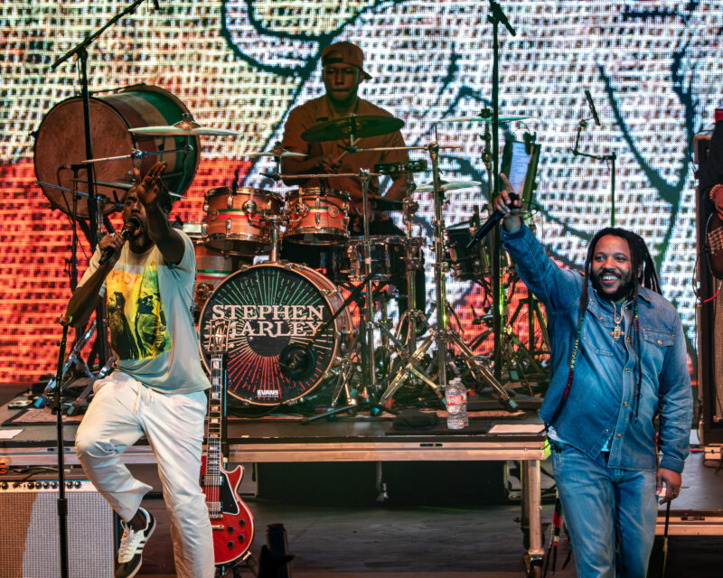 Singer front left with microphone on a brightly lit stage, drummer Stephen Marley in the center behind a drum kit, guitarist at left, and another performer in denim on the right; colorful LED backdrop fills the scene.
