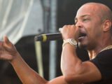 Male singer on stage, singing into a microphone with his left hand extended toward the audience.