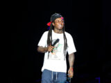 Male performer on stage, wearing a white graphic t-shirt and black cap backward, smiling while holding a microphone; dark background with spotlight.