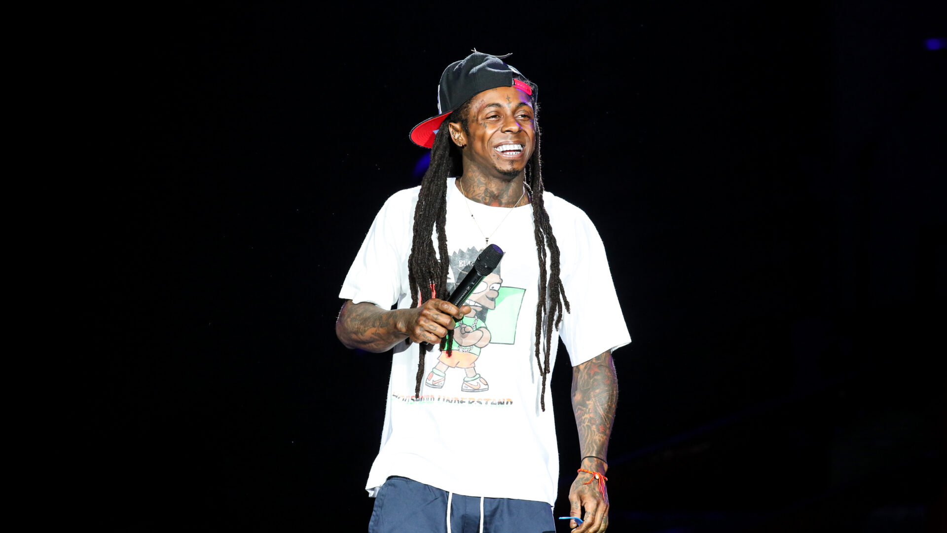 Raleigh,,Nc,-,July,27:,Lil,Wayne,Performs,In,Concert – The 824 Journal Male performer on stage, wearing a white graphic t-shirt and black cap backward, smiling while holding a microphone; dark background with spotlight.
