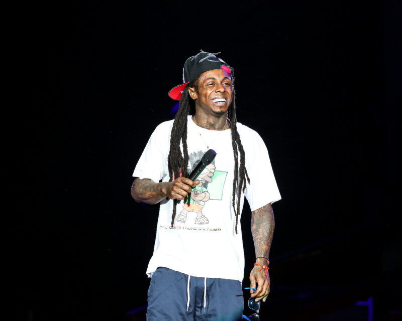 Male performer on stage, wearing a white graphic t-shirt and black cap backward, smiling while holding a microphone; dark background with spotlight.