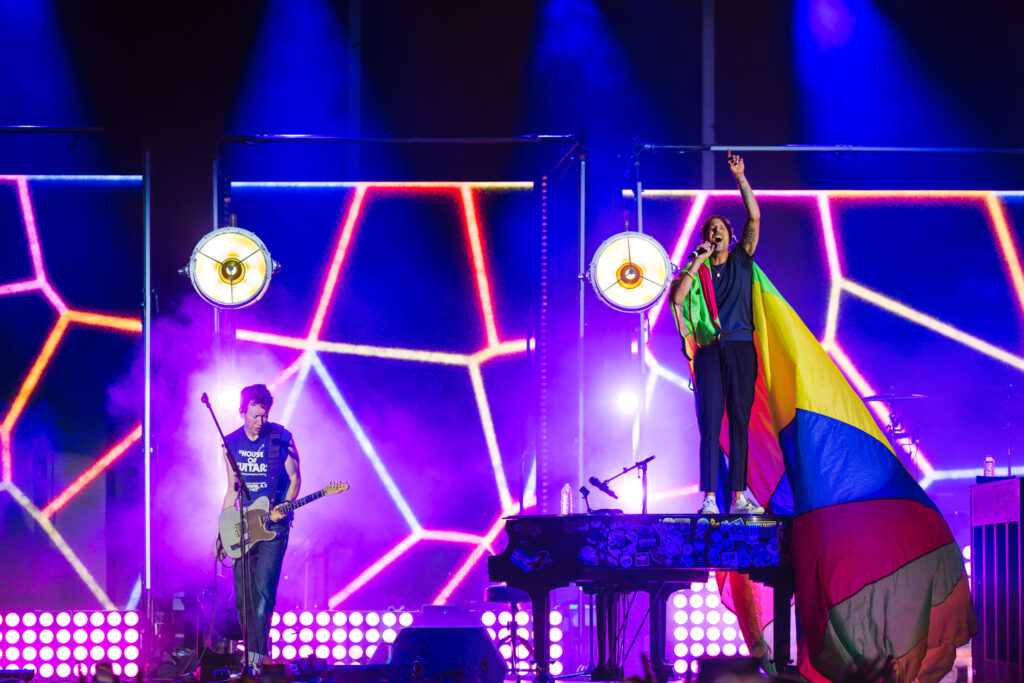 Male singer on stage wrapped in a rainbow flag, microphone in hand, raising his right arm amid neon geometric lights.