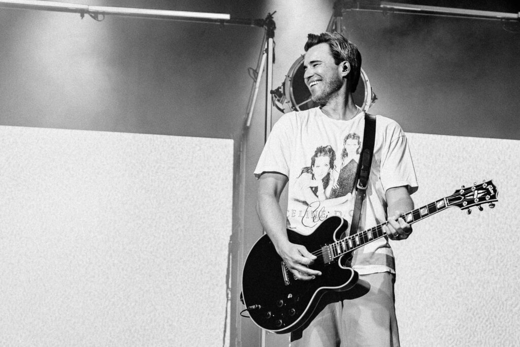 Guitarist on stage smiling while playing a black electric guitar in a white T‑shirt, spotlight behind him**