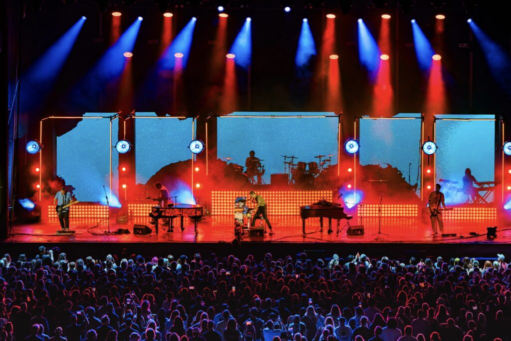 Live concert scene with a full band on stage under blue and red spotlights and a bright LED backdrop, crowd in the foreground.