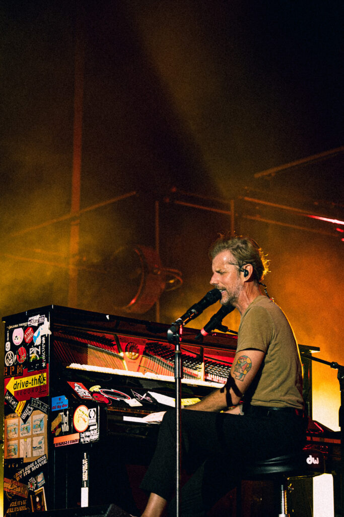 Male musician in an olive shirt sings into a microphone while playing a sticker-covered keyboard on a smoky, orange-lit stage.