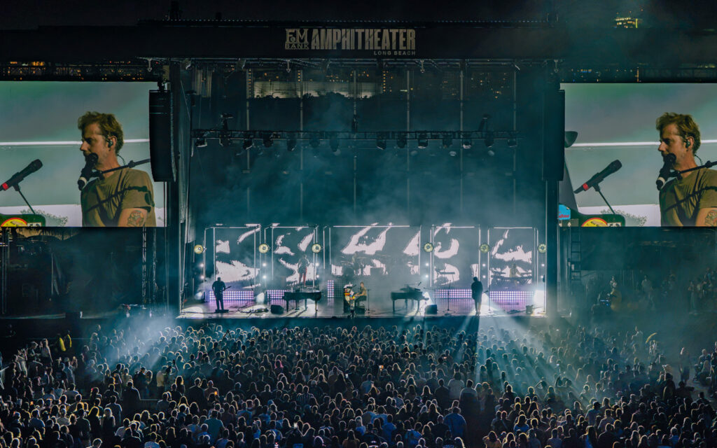 Large outdoor concert at night with a multi-level stage, side screens, performers, and a dense cheering crowd.