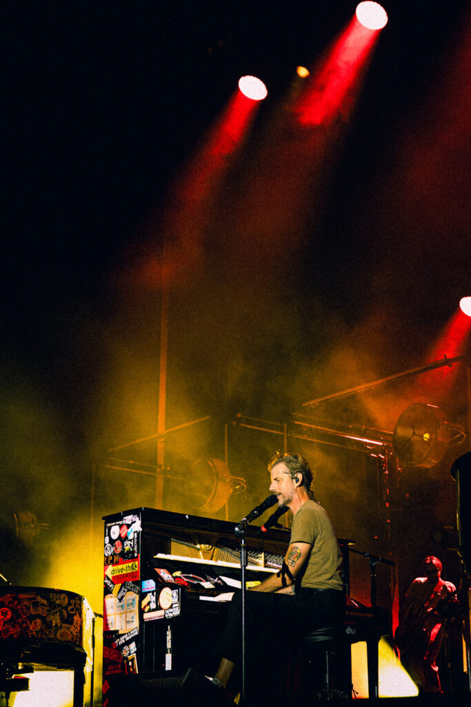 Male musician playing a sticker-covered keyboard on a smoky, yellow-lit stage with red spotlights.