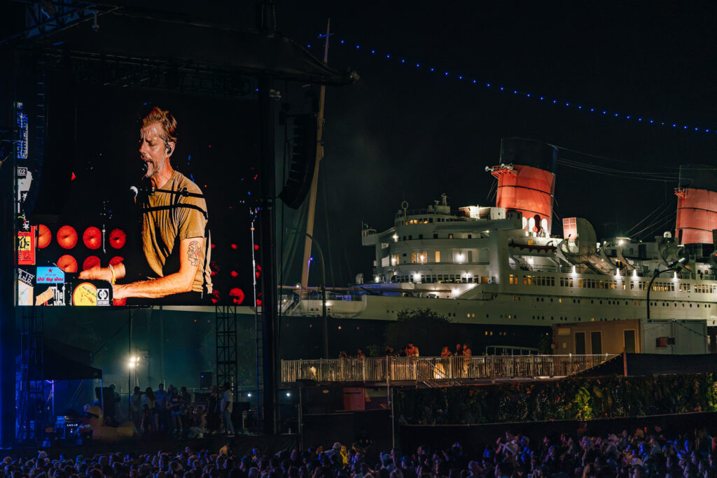 Nighttime outdoor concert on a cruise ship with a large screen showing a male singer performing, blue string lights above and a crowd in front.