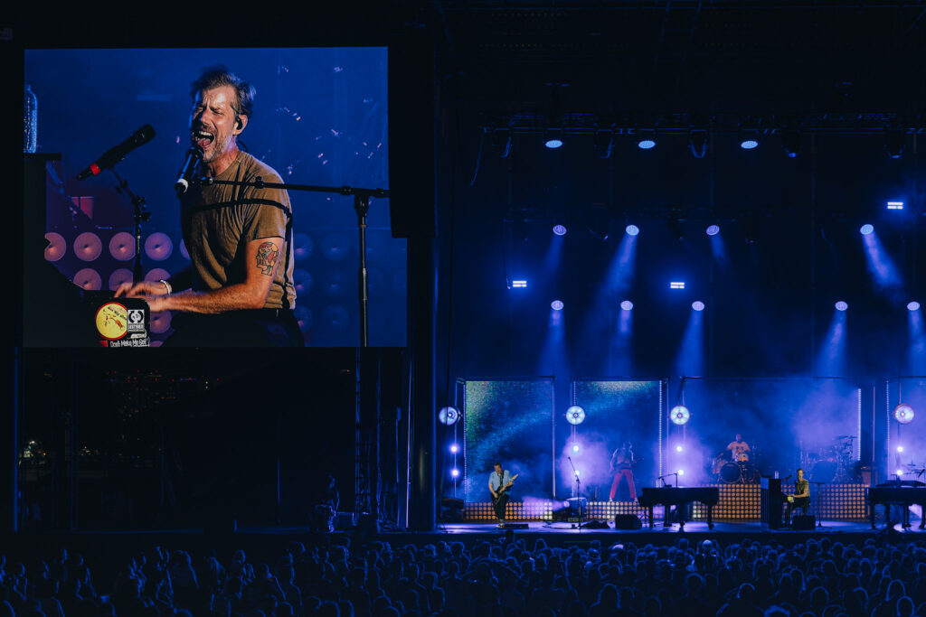 Singer performing on stage with a large screen projection to the left, wearing a tan shirt under blue lighting, audience in front.