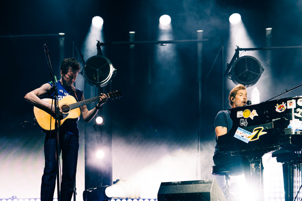 Two musicians perform on a smoky stage: a guitarist left plays a light brown acoustic guitar, while a keyboardist/singer on the right atop a decorated piano sings into a mic amid bright stage lights.