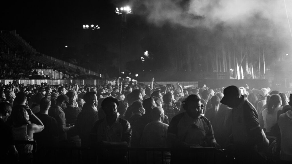 Crowd of concertgoers at night facing the stage, some hands raised, with fog and bright lights in the distance.