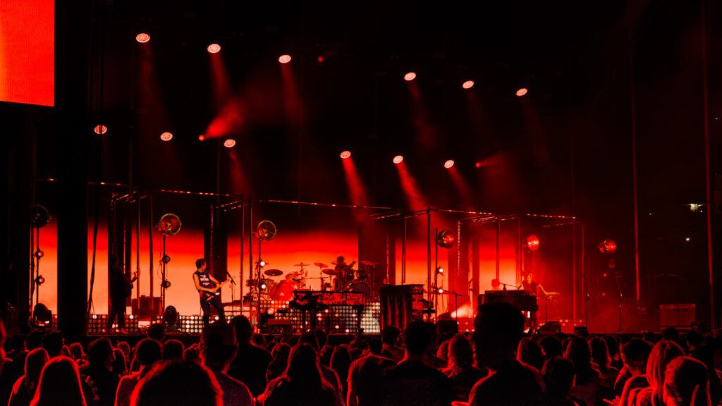 Live concert on a red-lit stage with musicians performing and a cheering audience in the foreground.