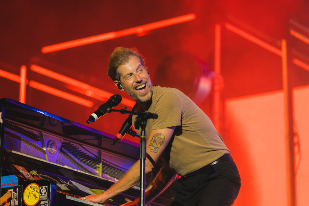 Male pianist on stage, smiling and singing into a microphone while playing a keyboard, red concert lighting behind him.