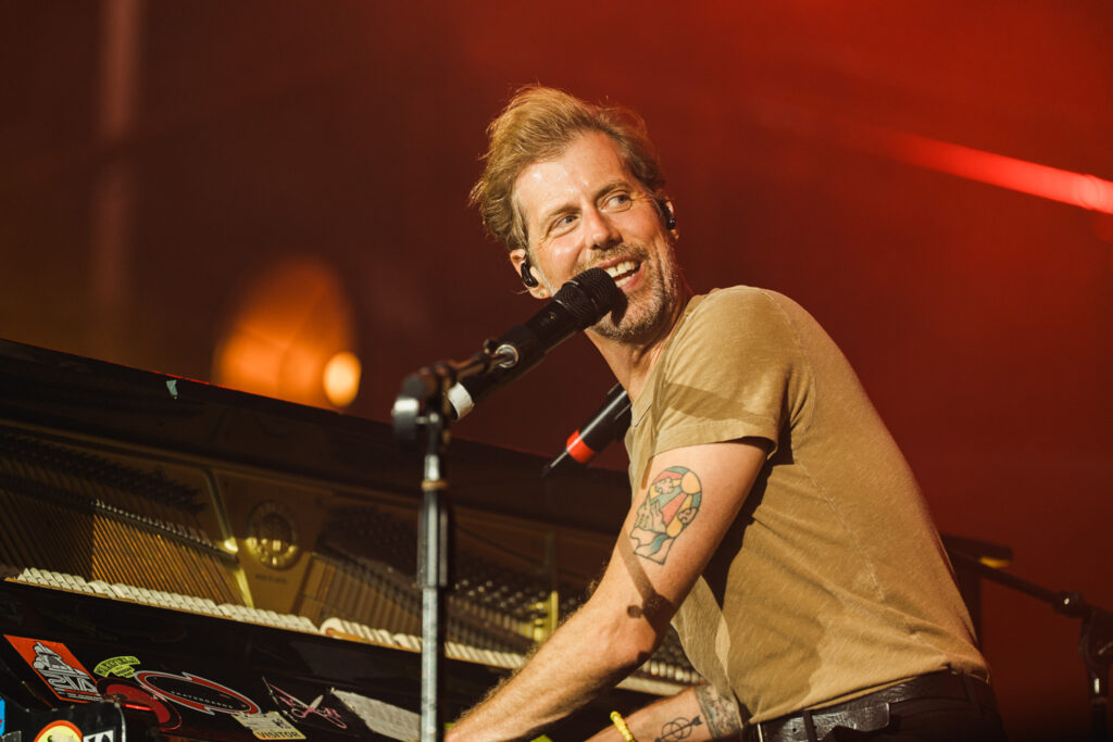 Male musician singing into a microphone while playing a grand piano on stage with red lighting.