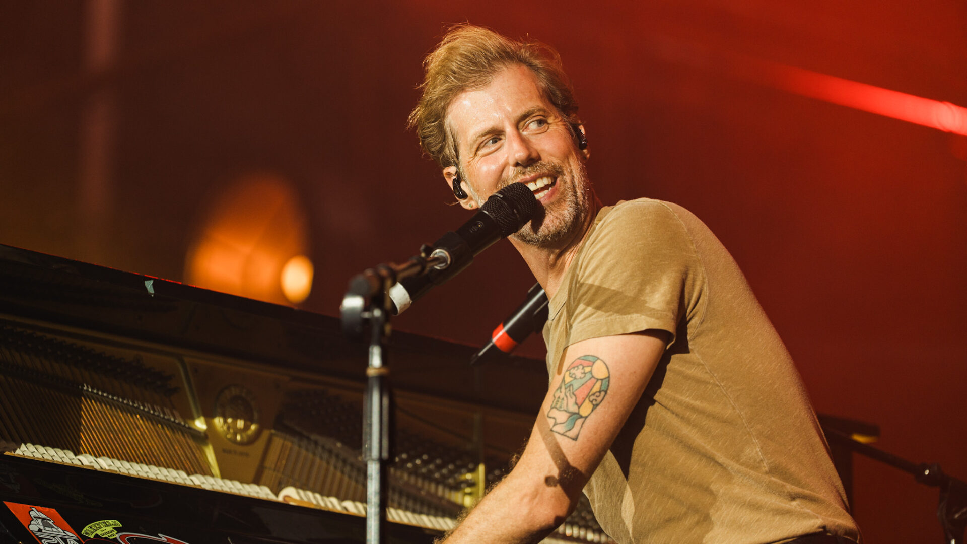 Male musician singing into a microphone while playing a grand piano on stage with red lighting.