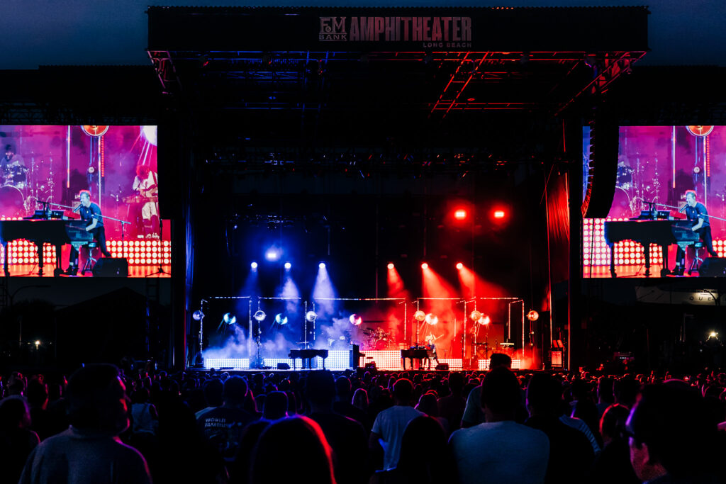 Nighttime outdoor concert with a large lit stage, blue and red lighting, side video screens showing performers, and a dense crowd in front.