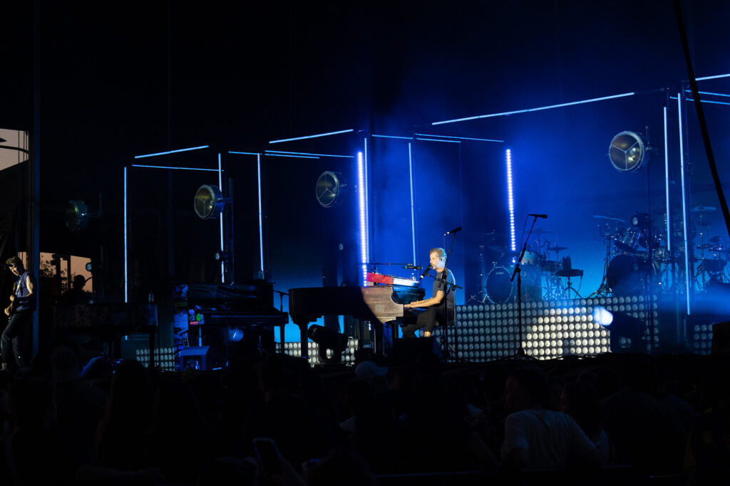 Musician playing a keyboard on a blue-lit concert stage with drums and equipment in the background.